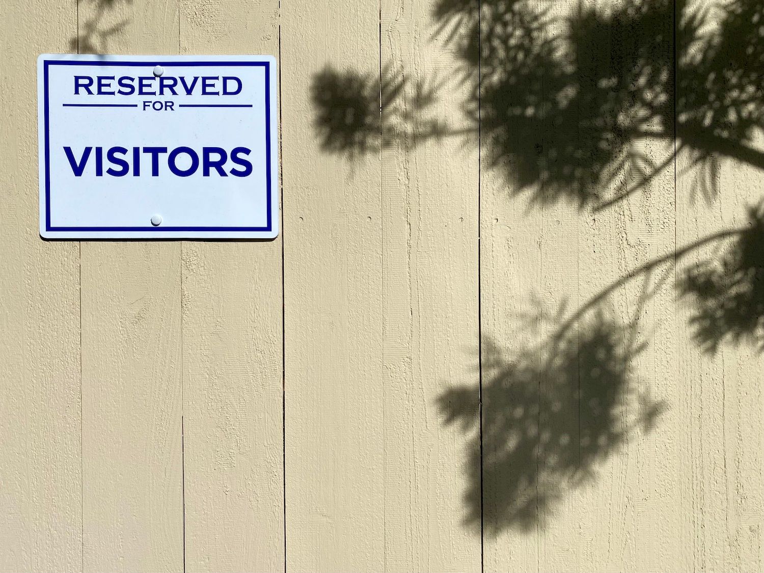 The shadow of a tree on a beige wooden fence, next to a white plastic sign with blue lettering that reads "RESERVED FOR VISITOR"