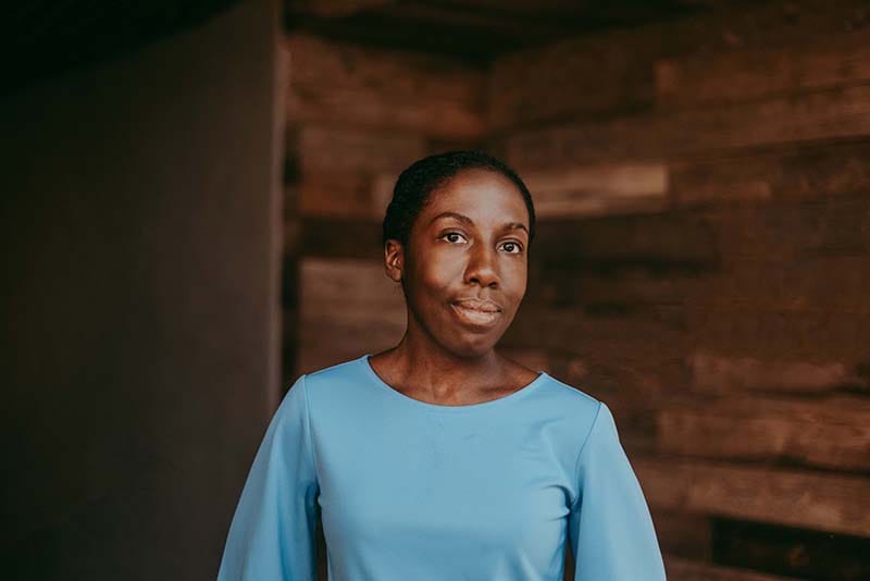 A portrait of writer Jennifer Baker. A black woman, centered in the frame in front of a wooden background, looks into the camera with a slight smile. She is wearing a blue shirt.