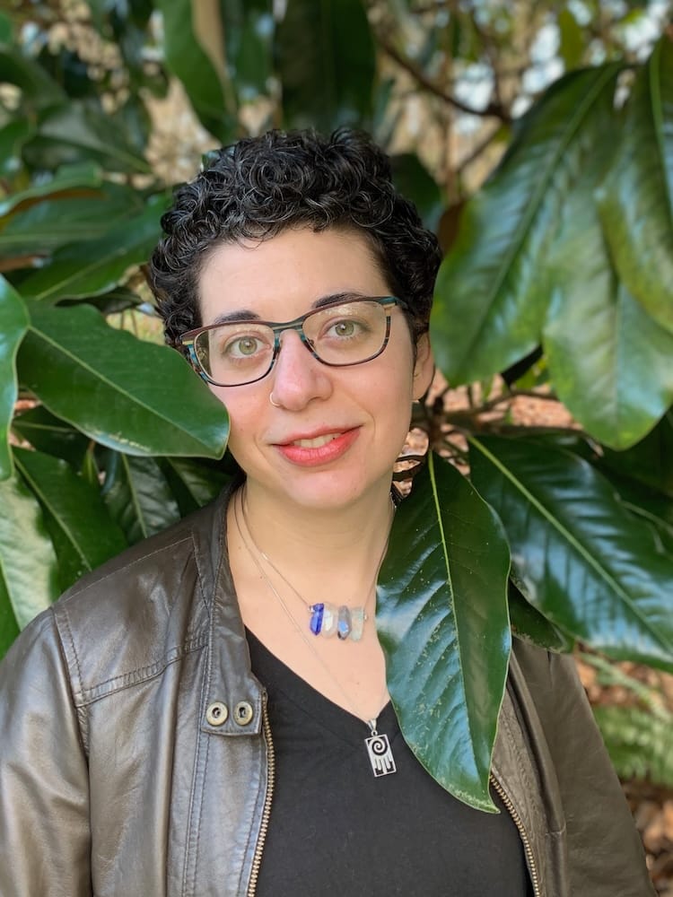 A portrait of poet Rachel Edelman. A white-presenting young woman looks into the camera. She has short, dark, curly hair, glasses, and a nose ring. She's wearing a brown leather jacket over a black v-neck t-shirt, and two necklaces, one with some crystals and one with a rectangular pendant.