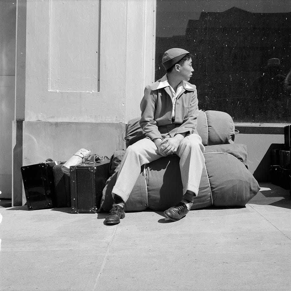 A black and white photograph of a Japanese American teenage boy. He is sitting on a large duffel bag tied with string, on the sidewalk next to some suitcases. He is looking out of frame over his left shoulder, his hands folded in his lap and with one foot cocked on its edges. His body language looks anxious to me.