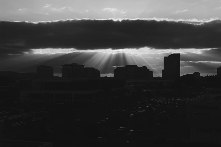 A high-contrast black-and-white photo of sunbeams shining from behind clouds onto a dark city skyline